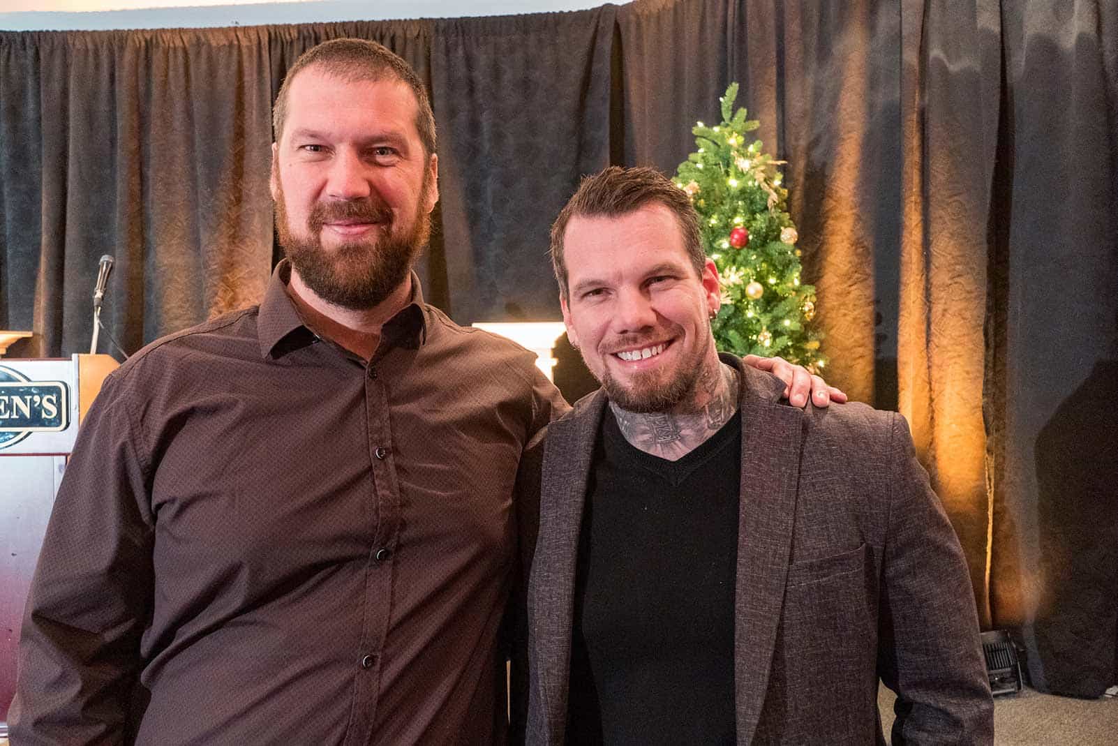 Two men pose beside each other for a picture, with a Christmas tree in the background.