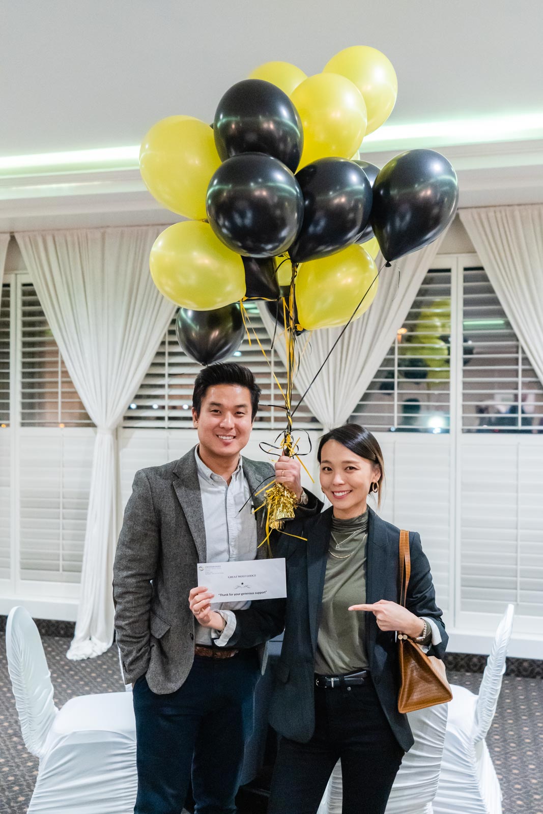A young man and woman smile and stand side by side in a reception lobby, holding an envelope and a large bundle of balloons.