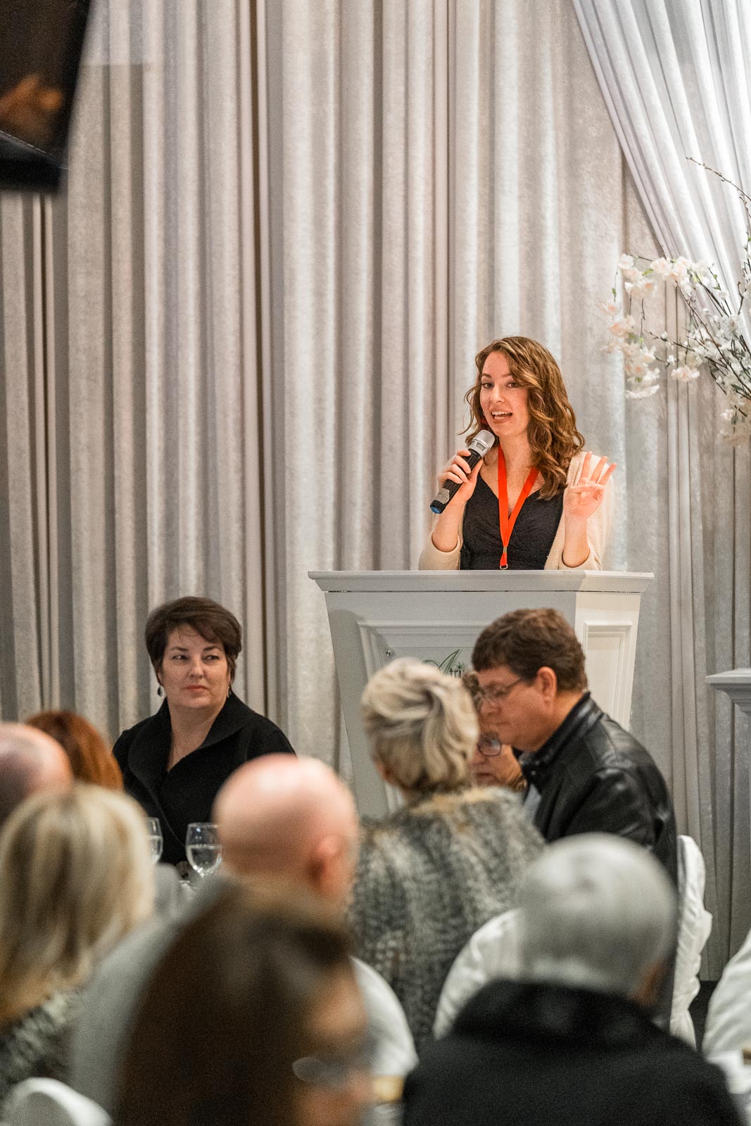 A woman stands at a podium, holding a mic and gesturing with her hands.
