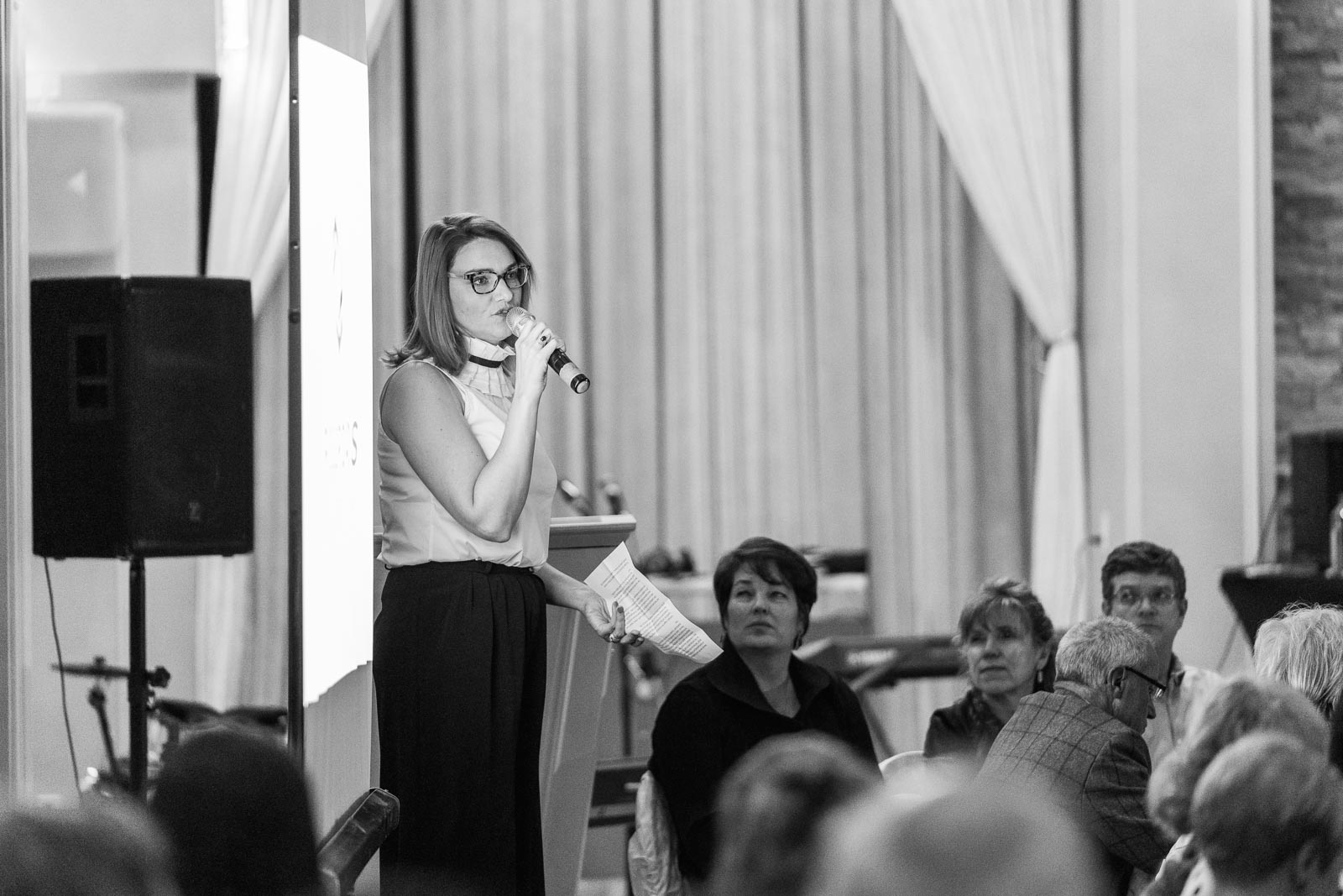 A black and white image of a woman standing on stage, holding a mic, making a presentation to a rapt audience. She's holding a sheet of paper in her other hand.