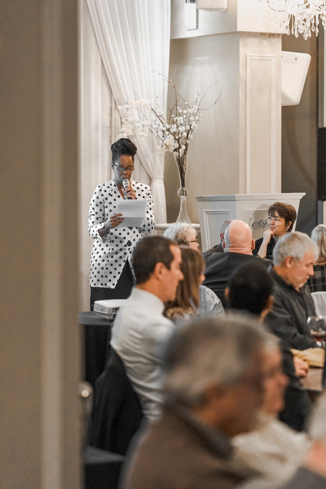 A tall woman reads speaks on stage, reading off a piece of paper.